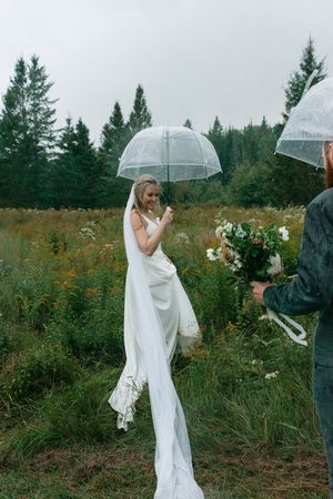 Person in flowing white dress holds clear umbrella while standing in rainy field with evergreen trees in background.
