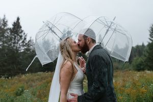 A romantic wedding kiss in the rain under clear umbrellas in a mountain meadow.
