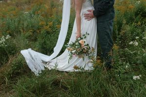 A flowing white wedding dress and bouquet of peach flowers against a backdrop of wild meadow grasses.