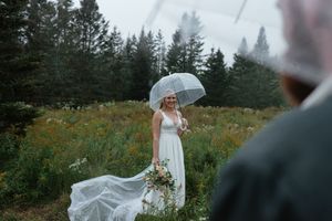 A bride in a flowing white dress holds an umbrella and bouquet while standing in a wildflower meadow near evergreen trees.