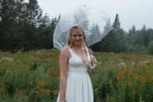 A person in a white dress stands in a wildflower meadow holding a clear umbrella with evergreen trees in the background.