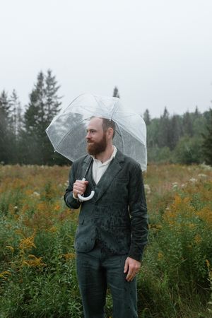 A suited figure holds a clear umbrella in a misty field of wildflowers.