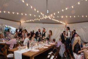 Wedding reception guests gather under a white tent decorated with string lights and wooden farm tables for dinner.