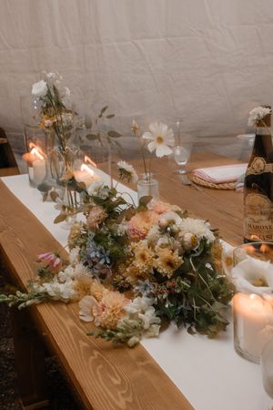Rustic wedding table centerpiece with white and blush flowers, greenery, and glowing candles on a white runner.