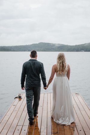 Couple walks hand in hand down a wooden dock overlooking a serene lake with mountains in the background.