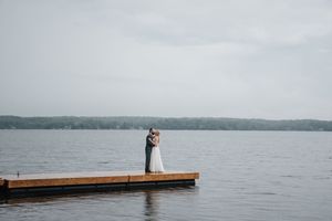 Two figures embrace on a wooden dock overlooking a serene lake surrounded by forested shoreline on an overcast day.