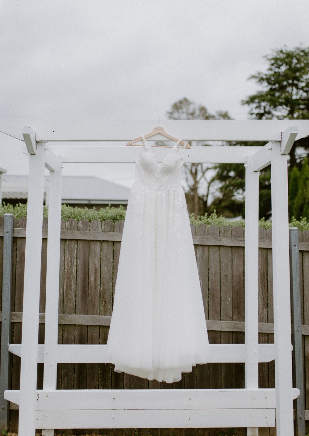White flower girl dresses hanging on a white wooden pergola against a wooden fence.