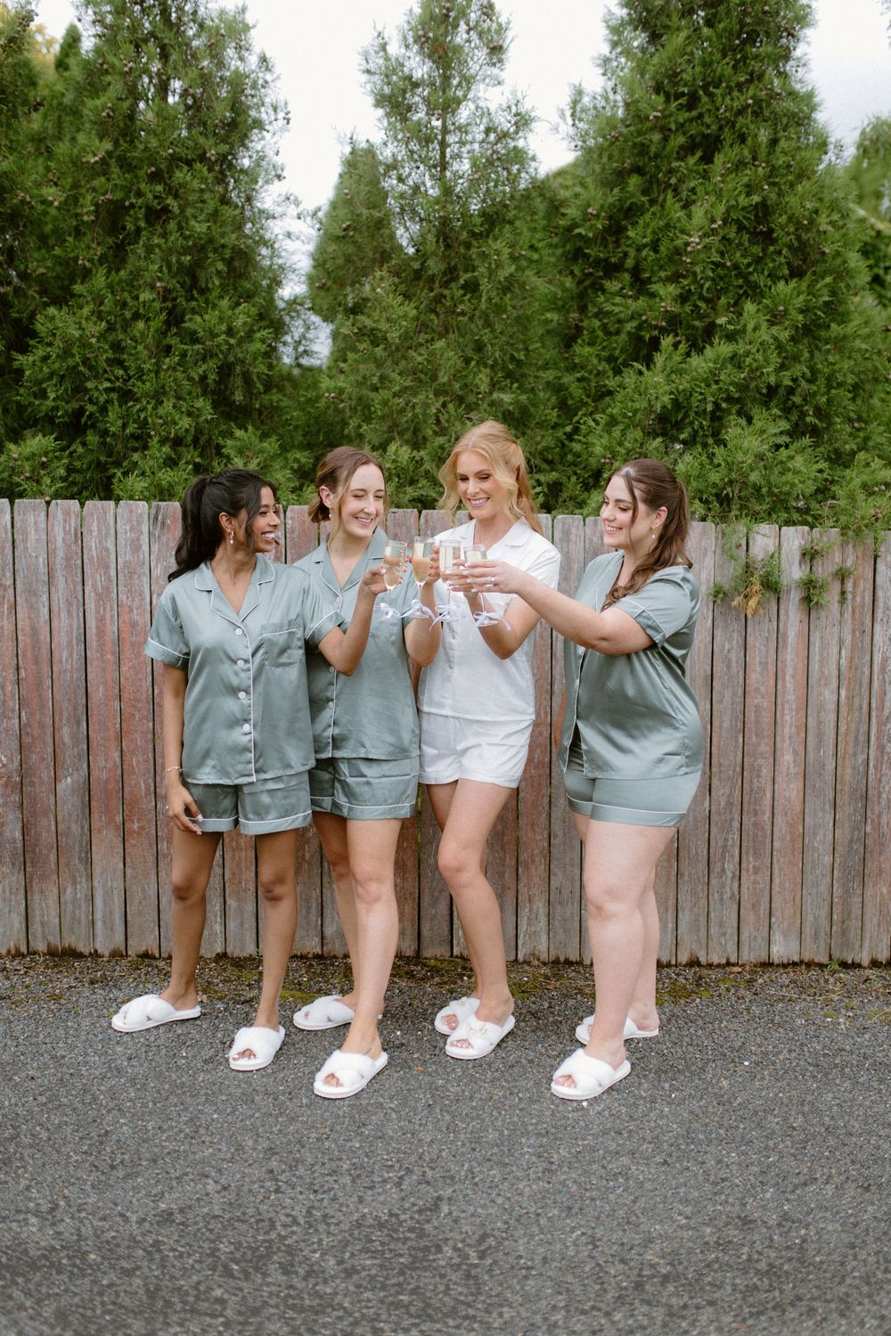 Bridesmaids in sage green robes posing playfully against a wooden fence.
