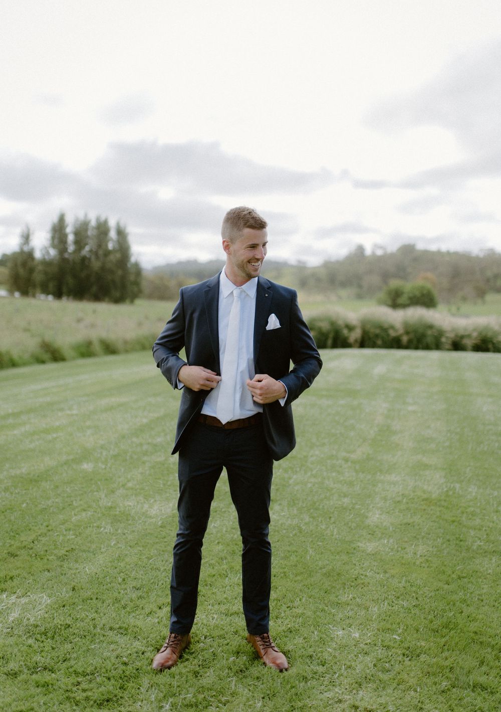 Groom in a dark suit poses playfully on a green lawn during wedding portraits.