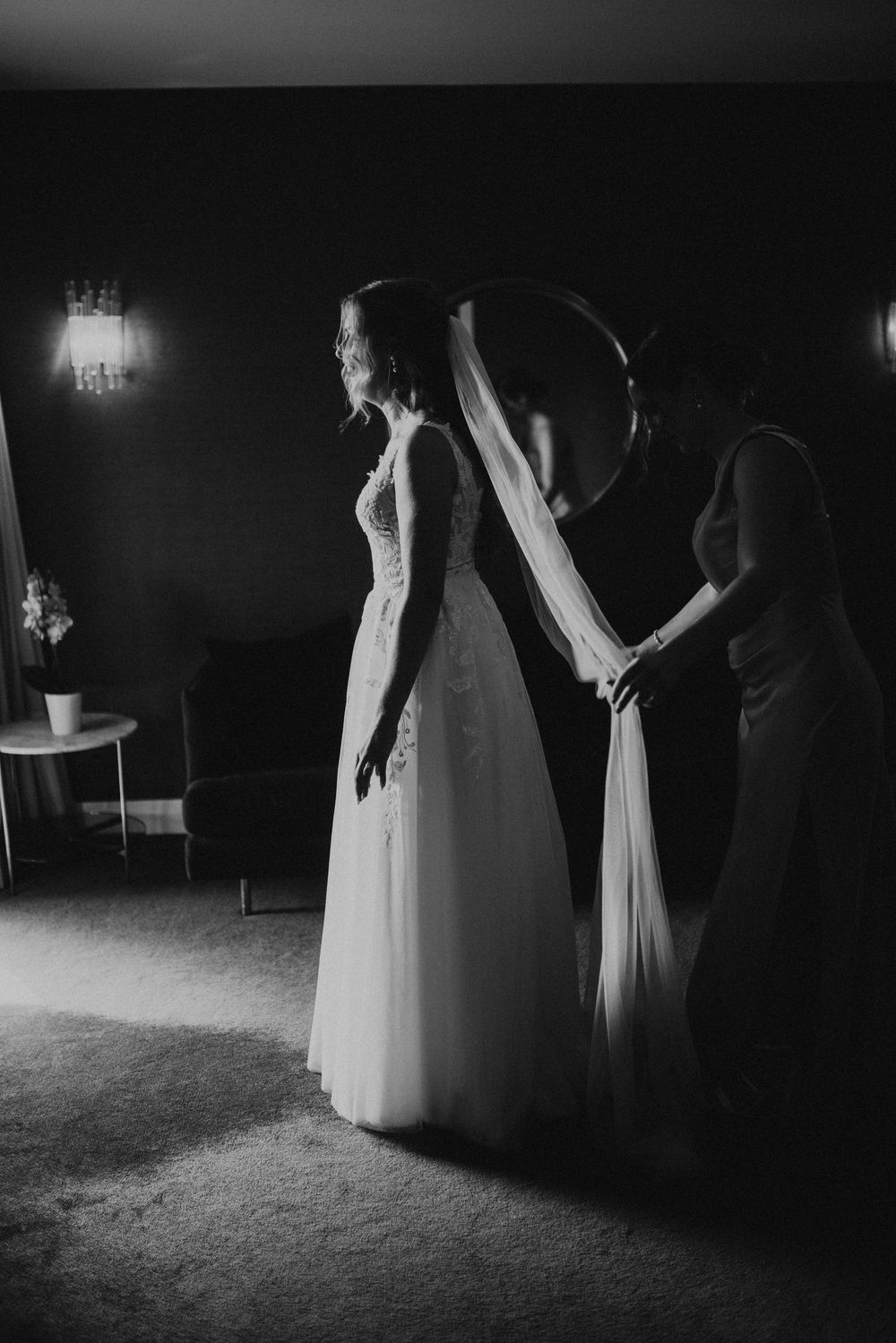 A black and white photo shows the final touches being made to a flowing white wedding dress in a dimly lit room.