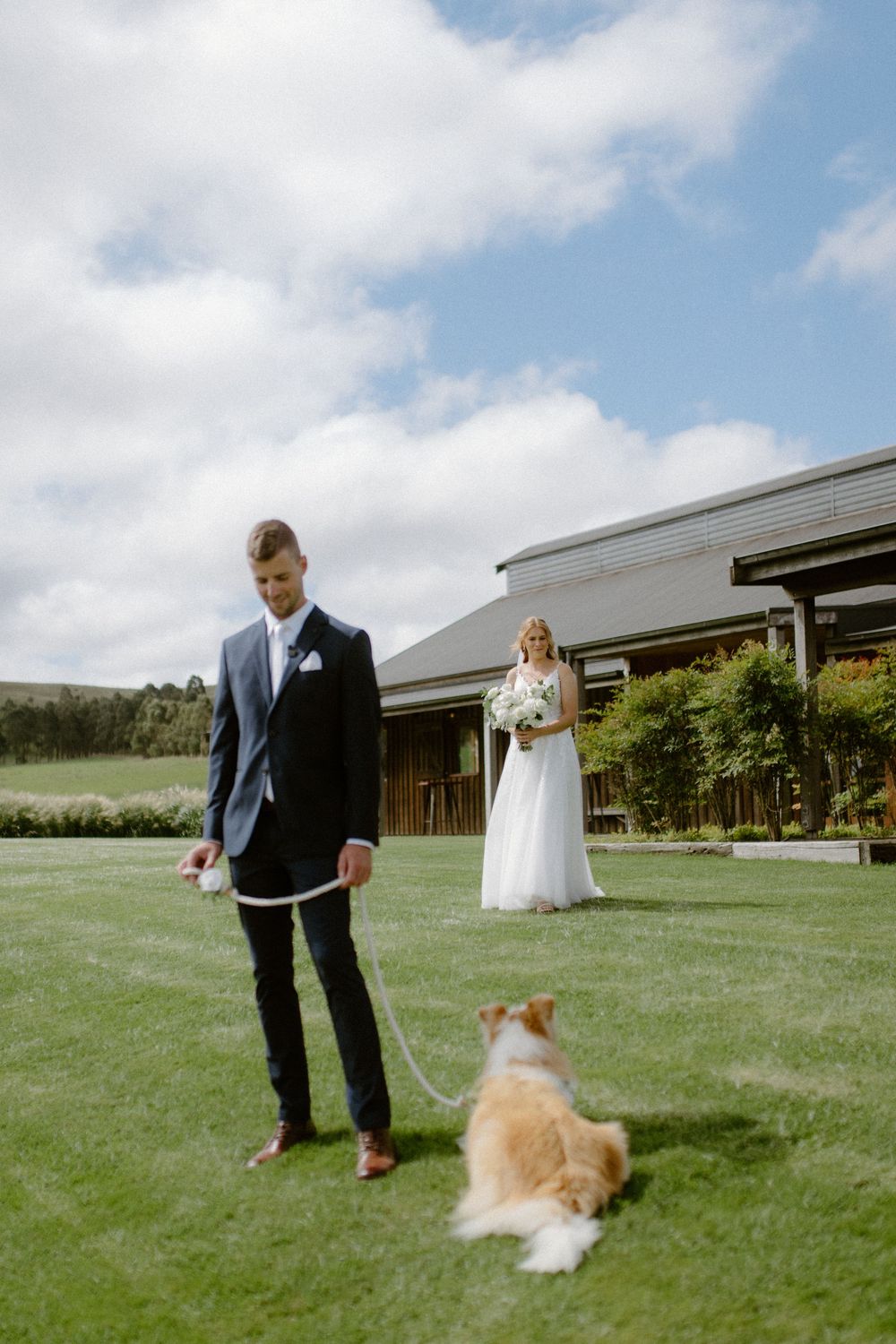 A wedding scene on a green lawn with the couple and their fluffy dog by a rustic venue.