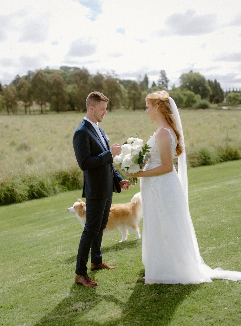 A couple exchanges wedding bouquets on a grassy hillside while their dog watches nearby.