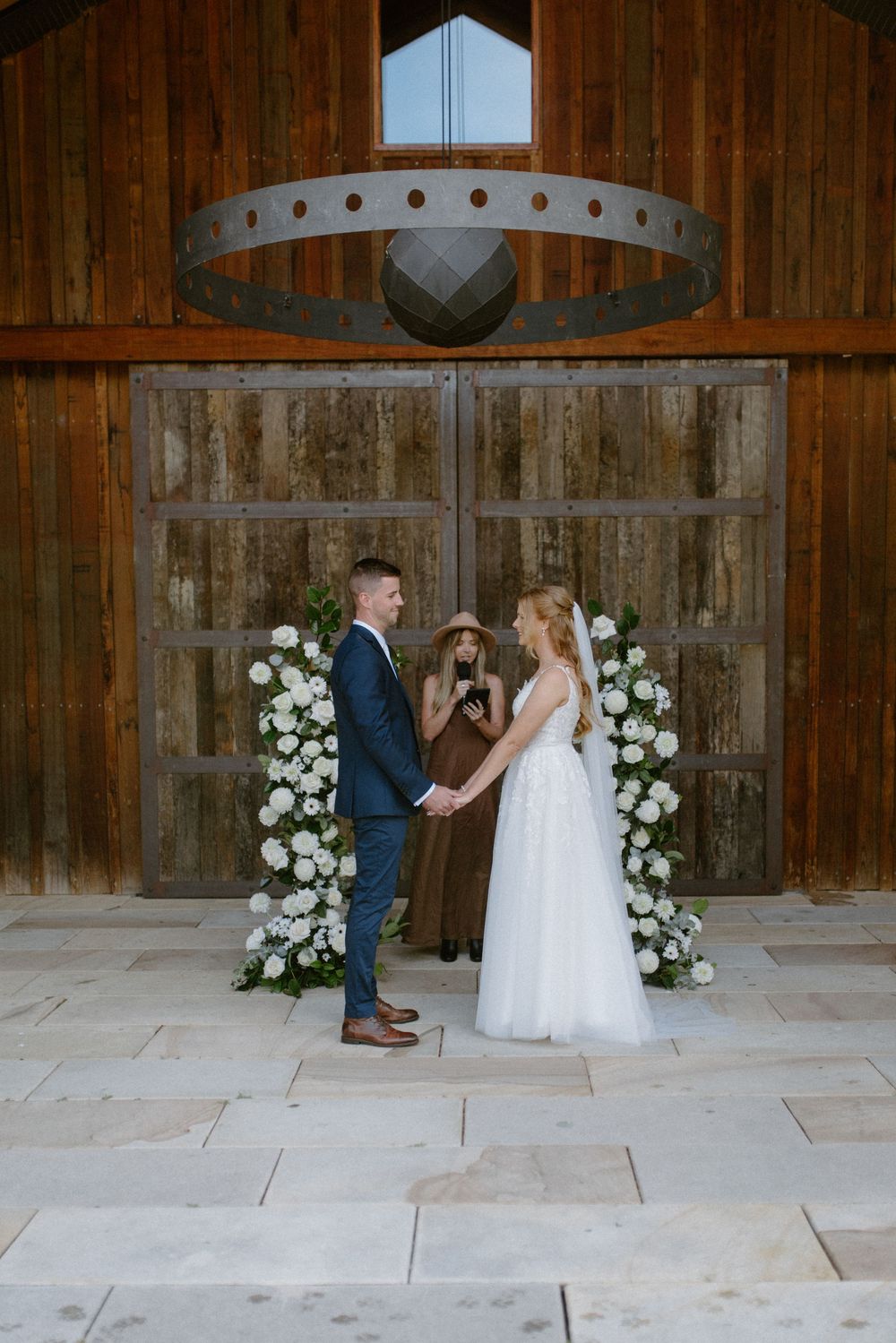 A wedding ceremony inside a rustic wooden barn with white floral arrangements framing the couple.