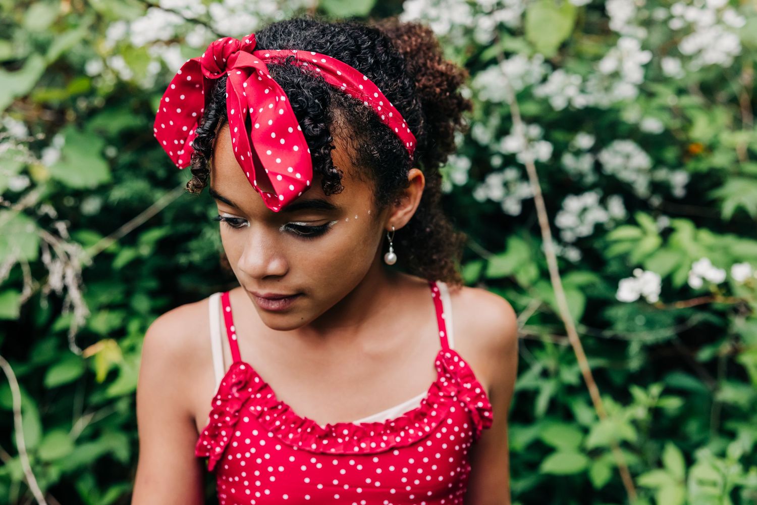 A girl in a red polka dot top and matching headband sits peacefully among green foliage and white flowers in a photo session at Brooksvale
