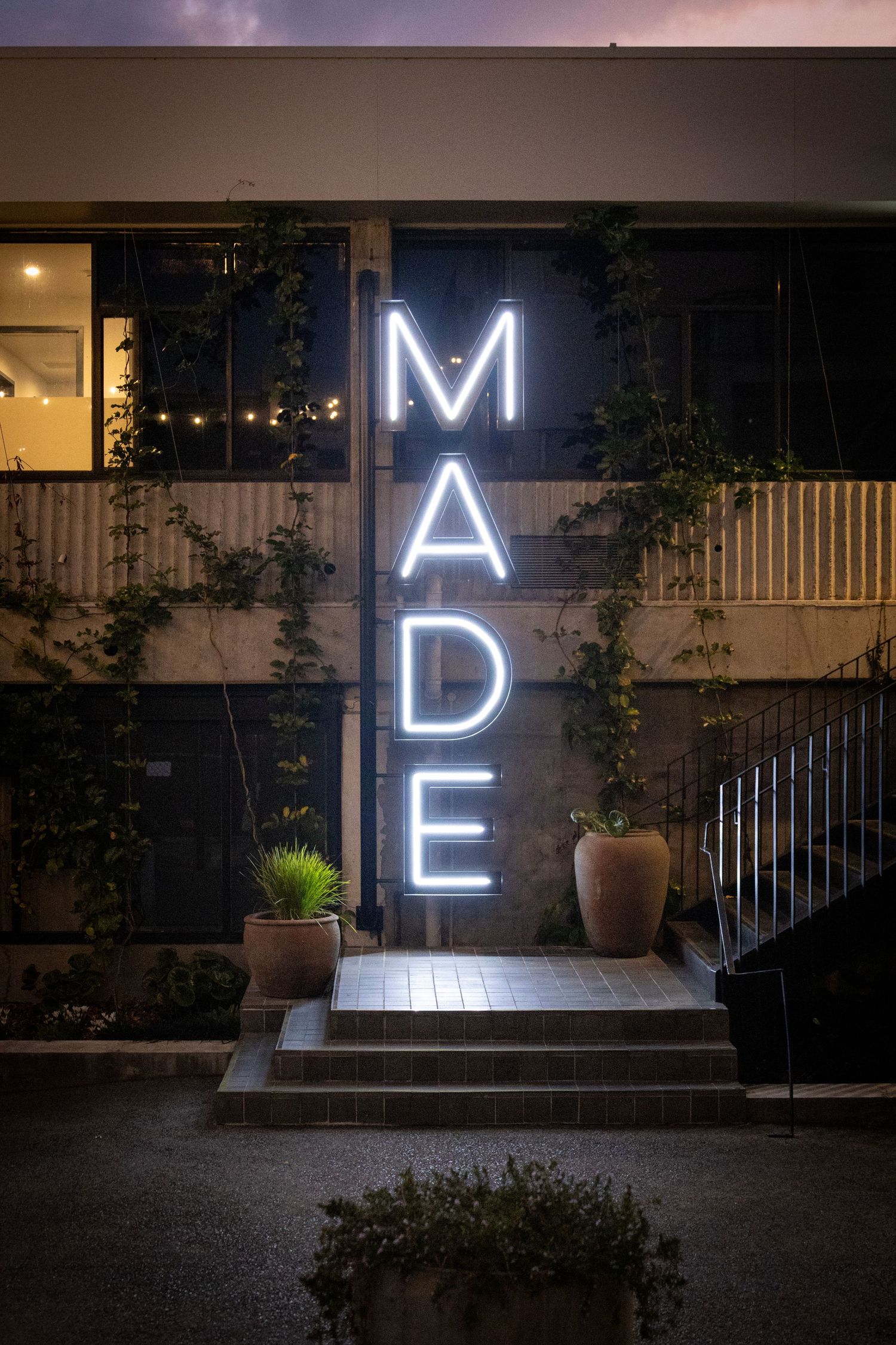 Illuminated “MADE” sign on a modern exterior with warm lights and potted plants at night.