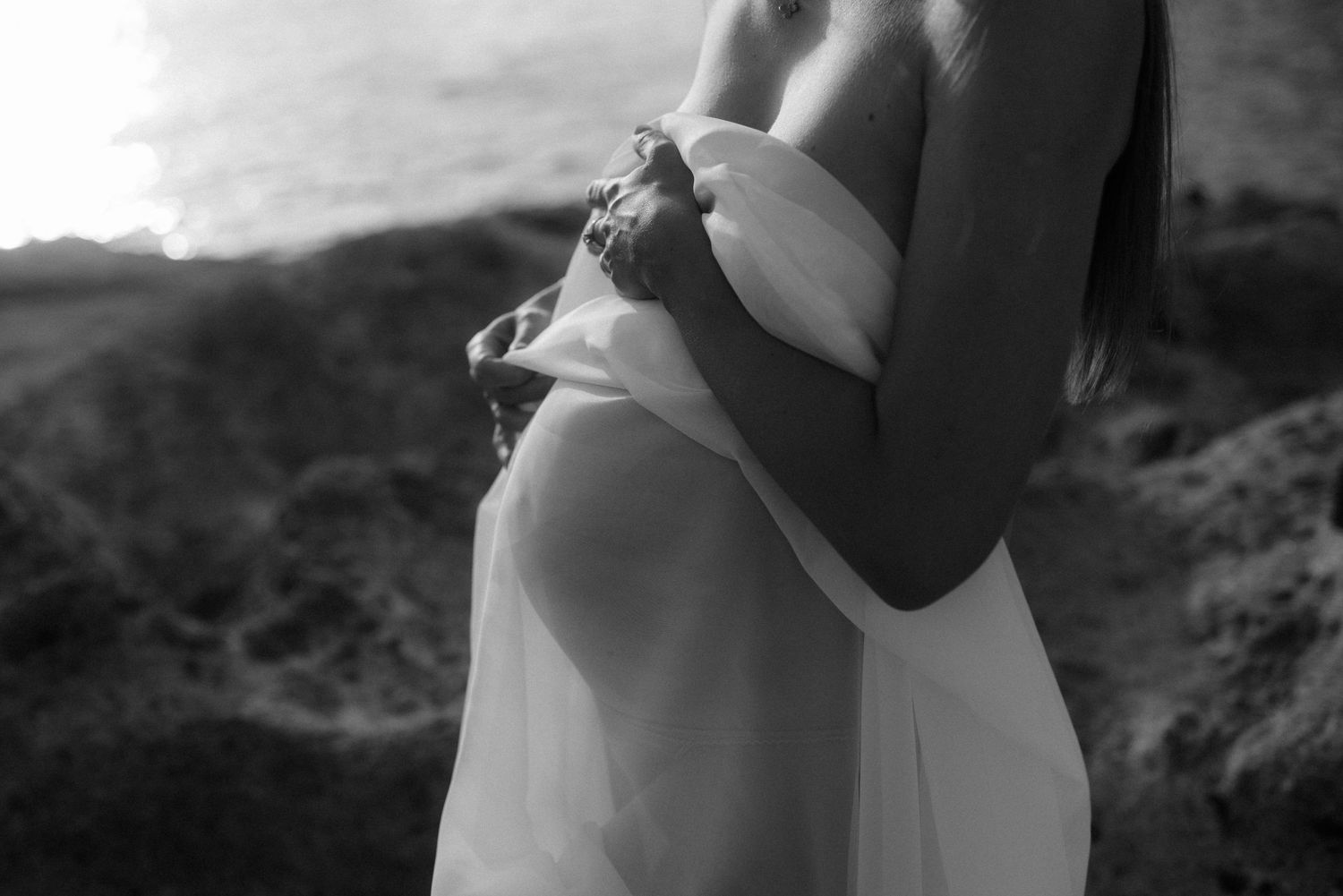 Black and white intimate portrait of expecting parents embracing outdoors with mother's pregnant belly visible.
