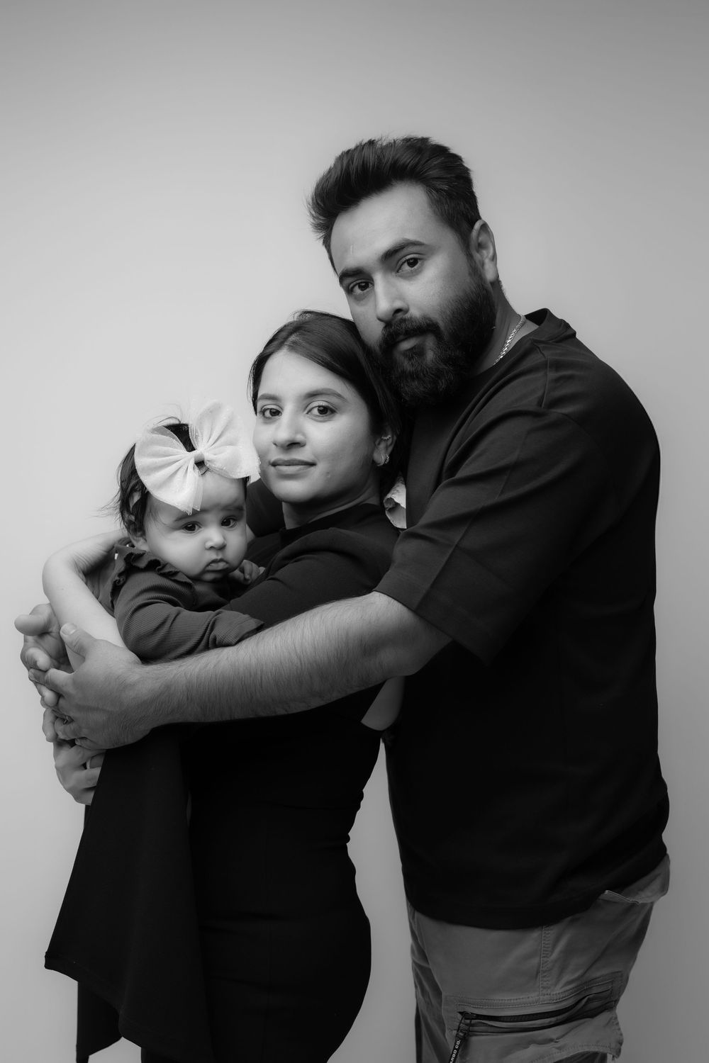 Black and white portrait of a family embracing together, featuring a baby with a bow in their hair.