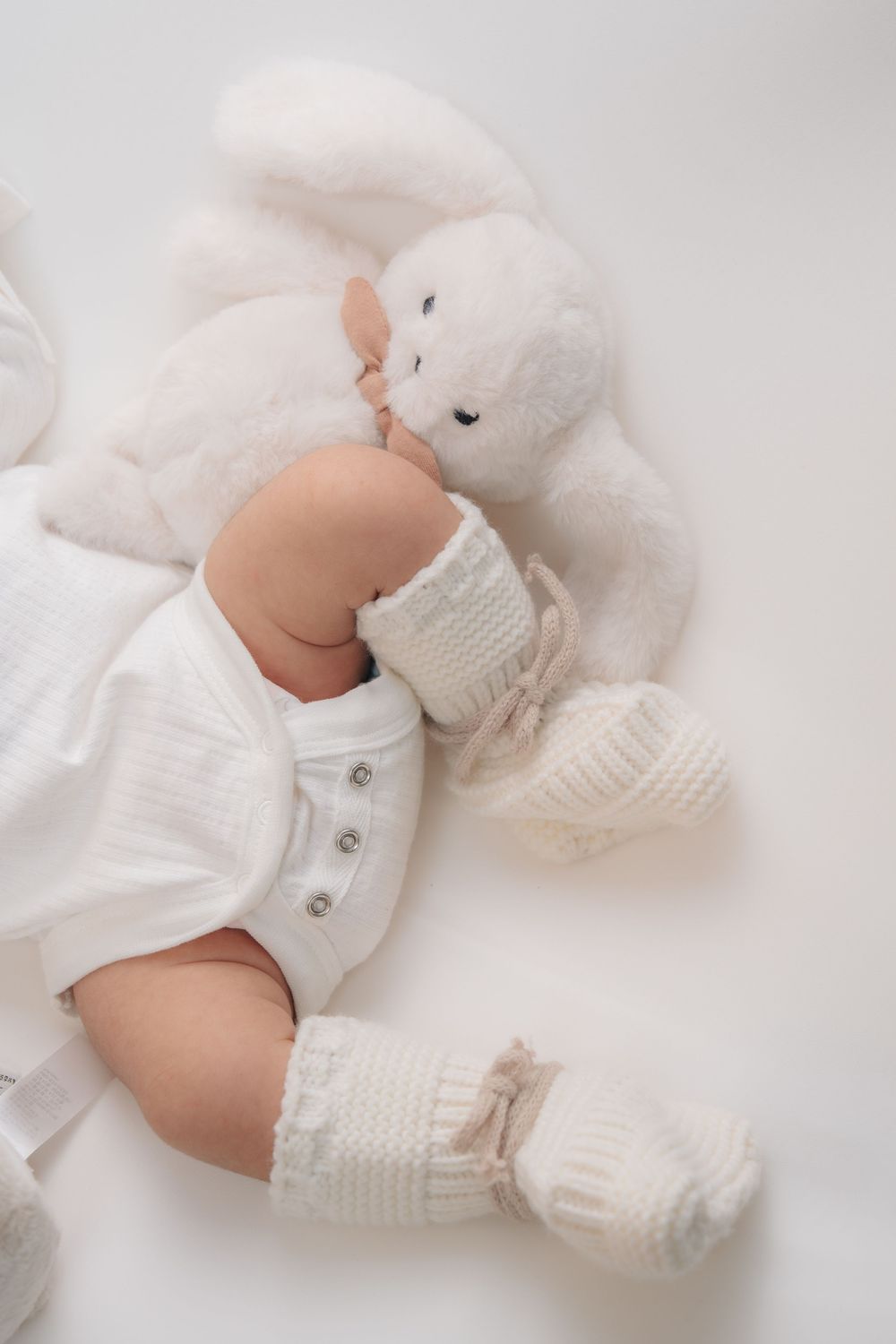 Baby wearing white knit clothing and socks lying next to a soft white teddy bear on white background.