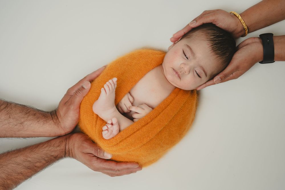 Newborn baby sleeping in orange bowl held by parents hands on white background.