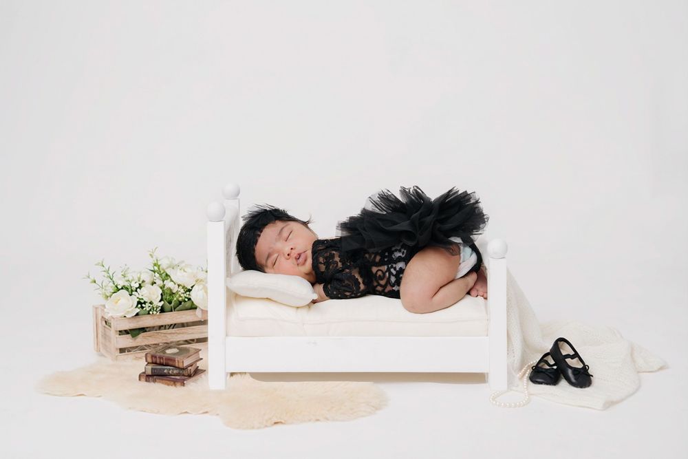 A sleeping baby in a black tutu dress lies on a white wooden bed prop with flowers and shoes nearby.