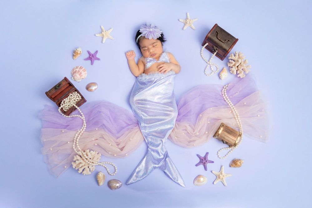 Newborn baby wrapped in silver swaddle surrounded by purple tulle, books, and starfish decorations in mermaid theme setup.