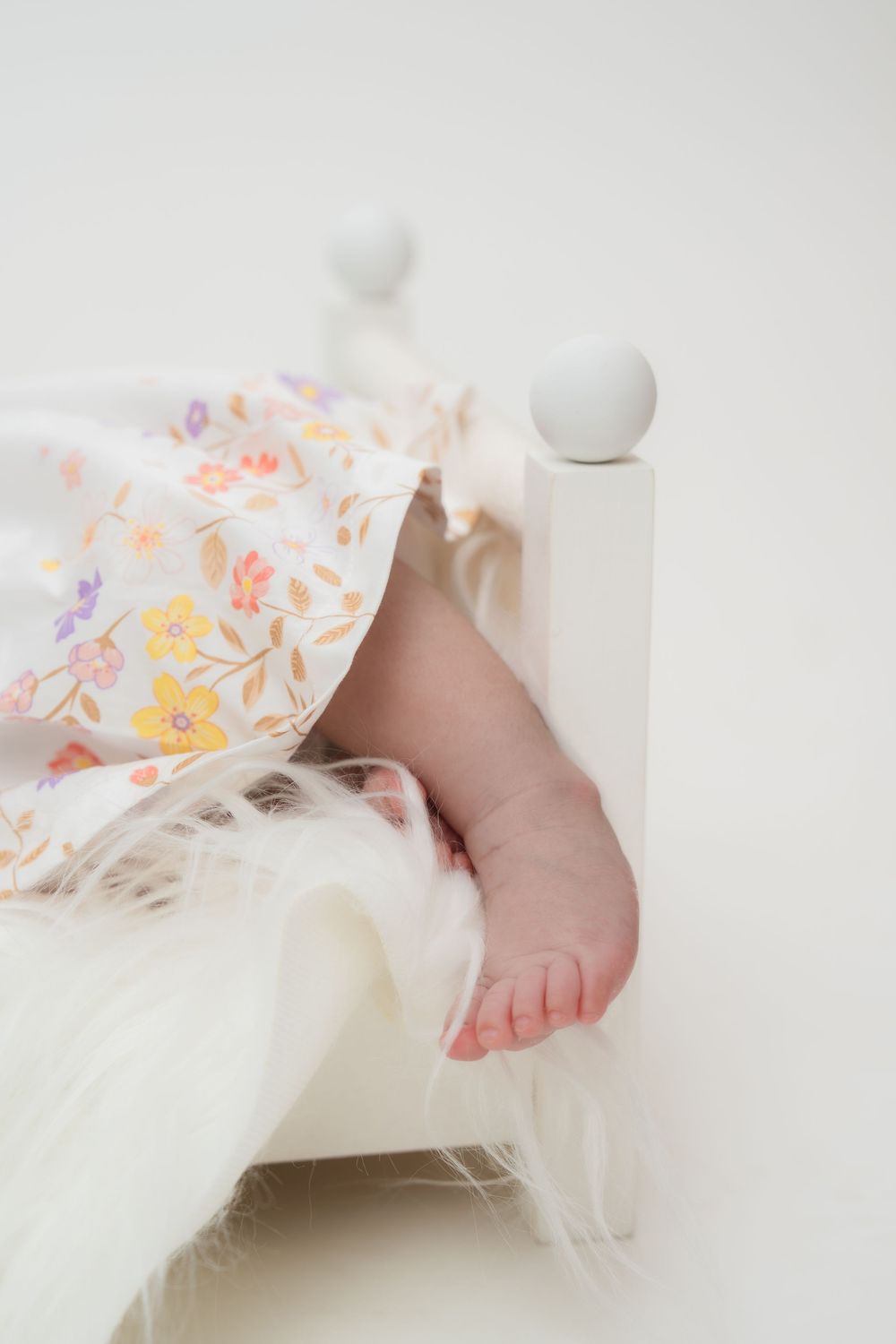 A tiny baby foot peeks out from under delicate white fabric with soft floral patterns in peach and yellow tones.