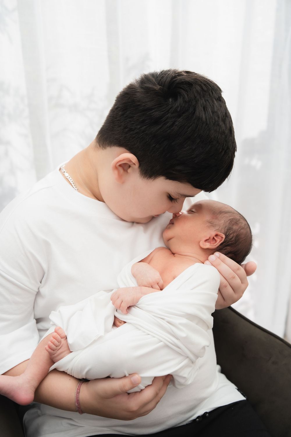 A tender moment captured in a series of photos showing a sibling gently kissing a baby on a white couch.