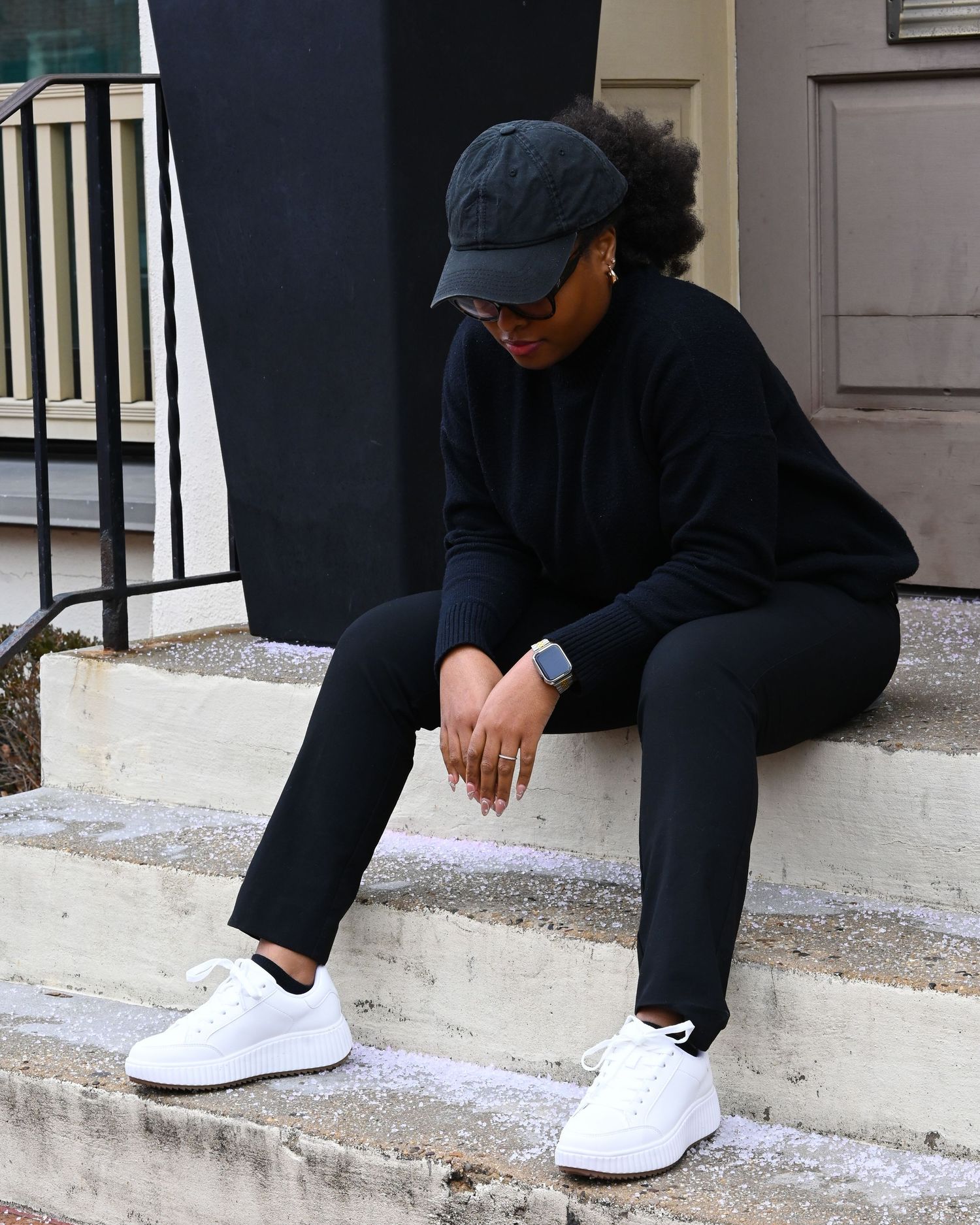 A figure wearing dark clothing and a cap sits pensively on concrete steps while wearing crisp white sneakers.