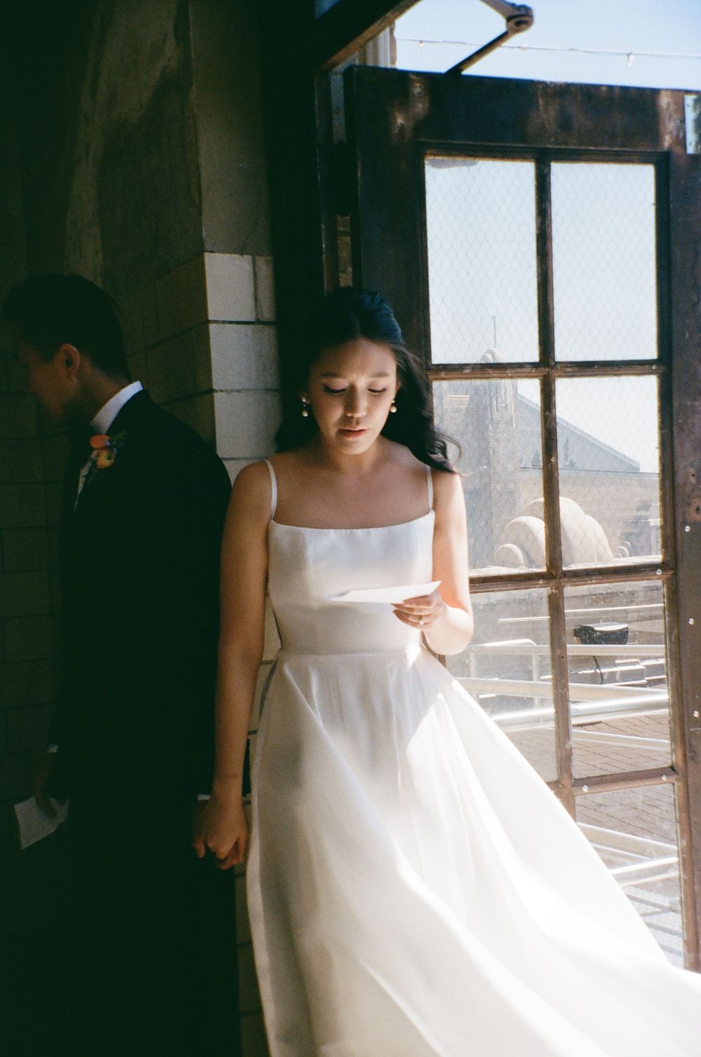 A person in a white wedding dress stands near a window in dramatic lighting.