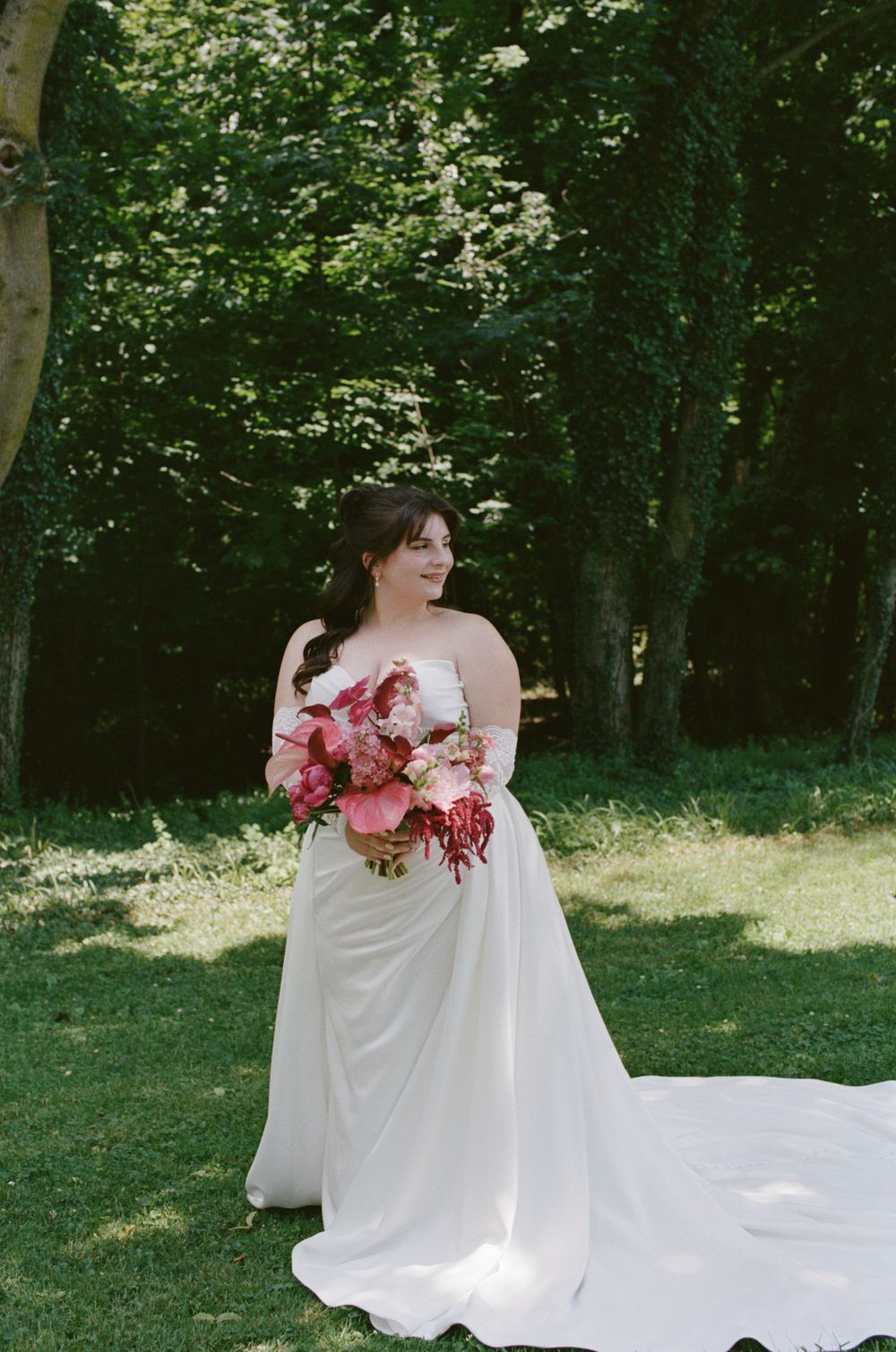 Bride in white strapless gown with long train holds pink floral bouquet outdoors among lush green trees.