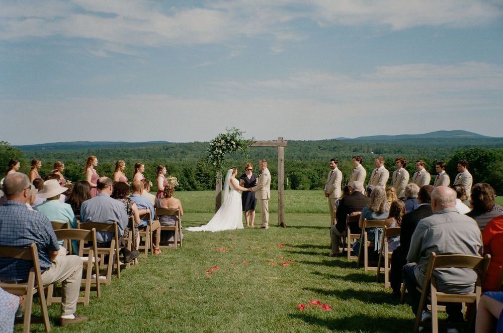 Panoramic view of an outdoor wedding ceremony in a scenic green field with rolling hills in the background.