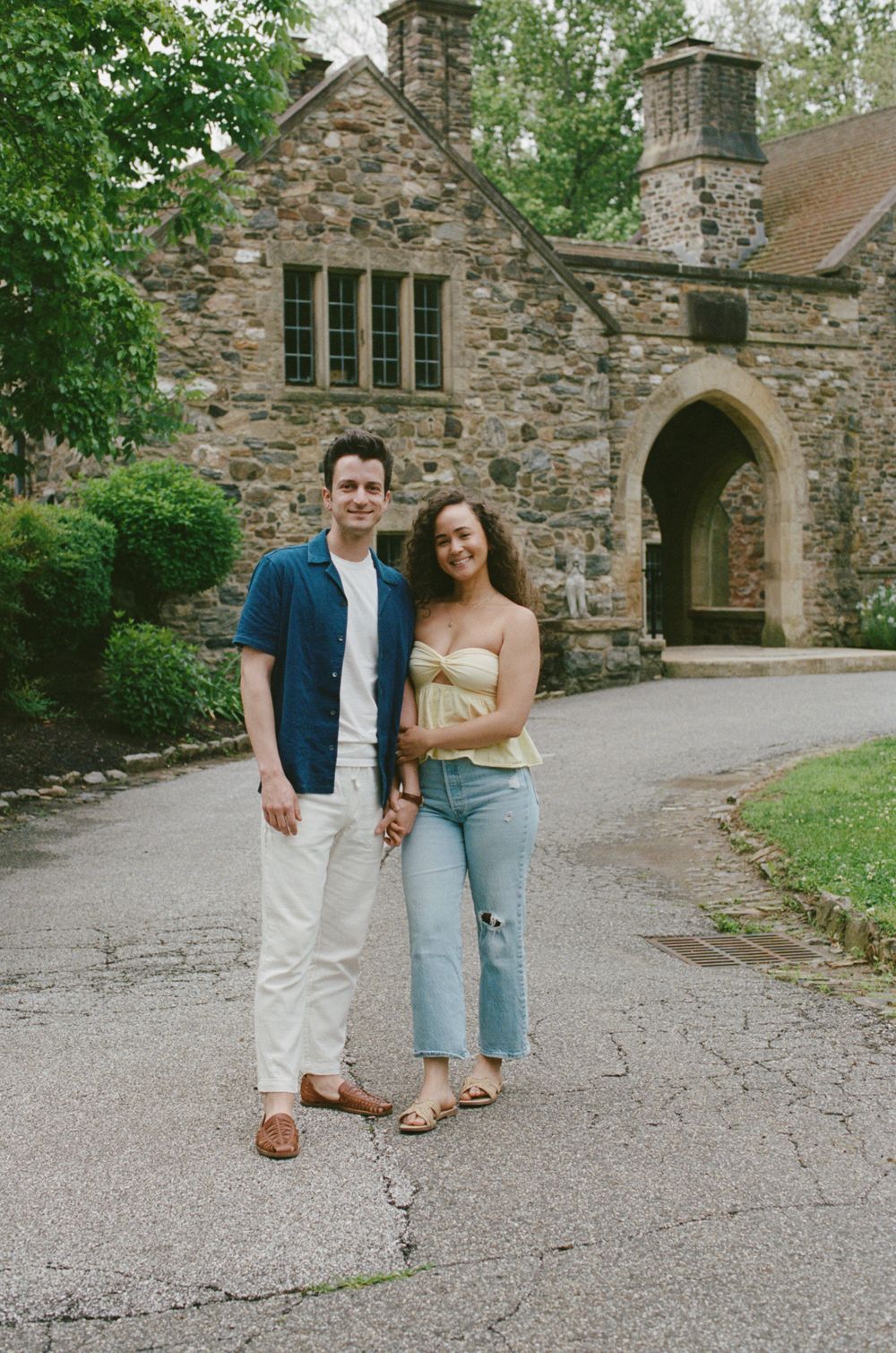 A couple smiles together in front of a historic stone building with an arched entrance on a tree-lined path.