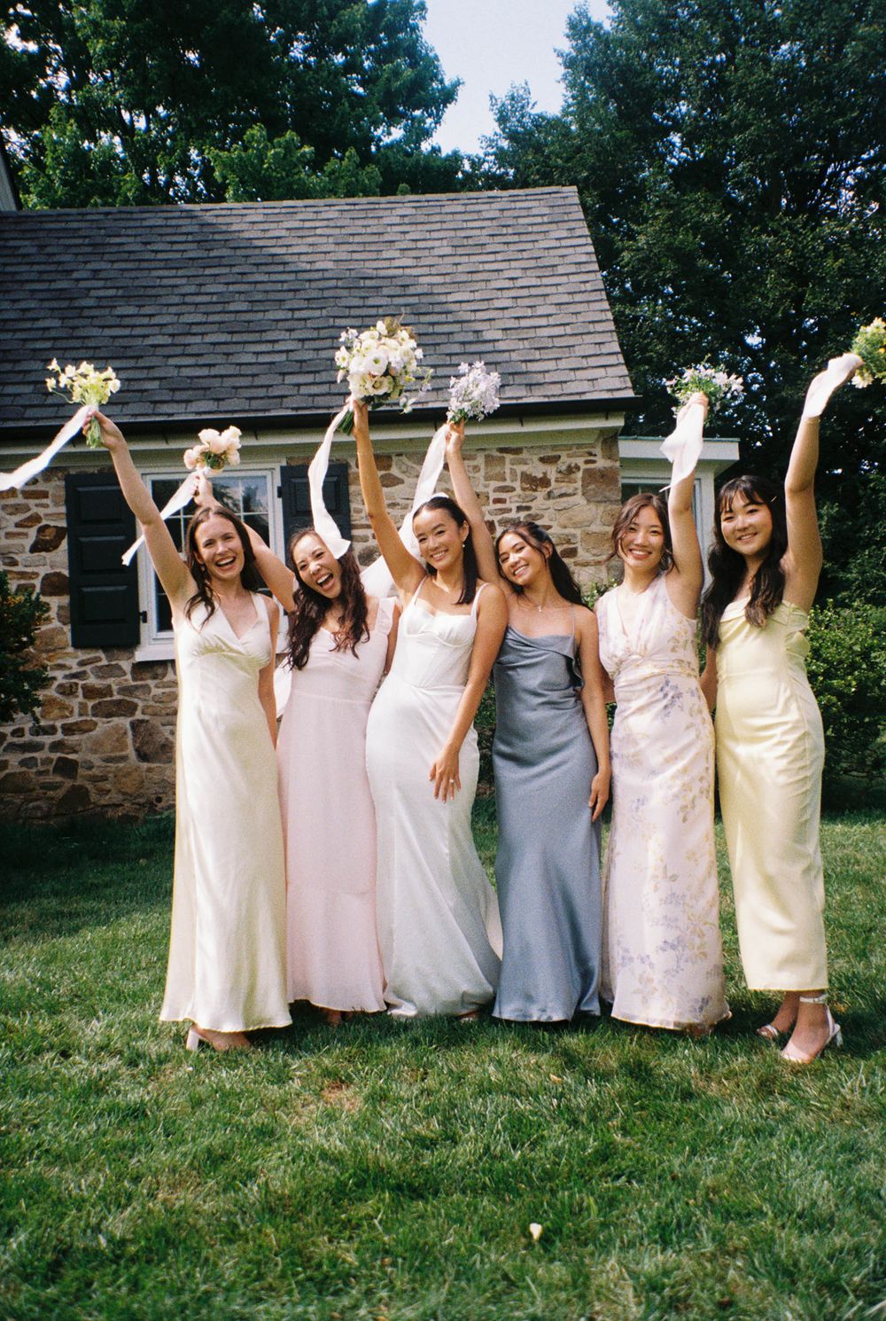 Bridesmaids in light colored dresses posing together on grass in front of stone building.