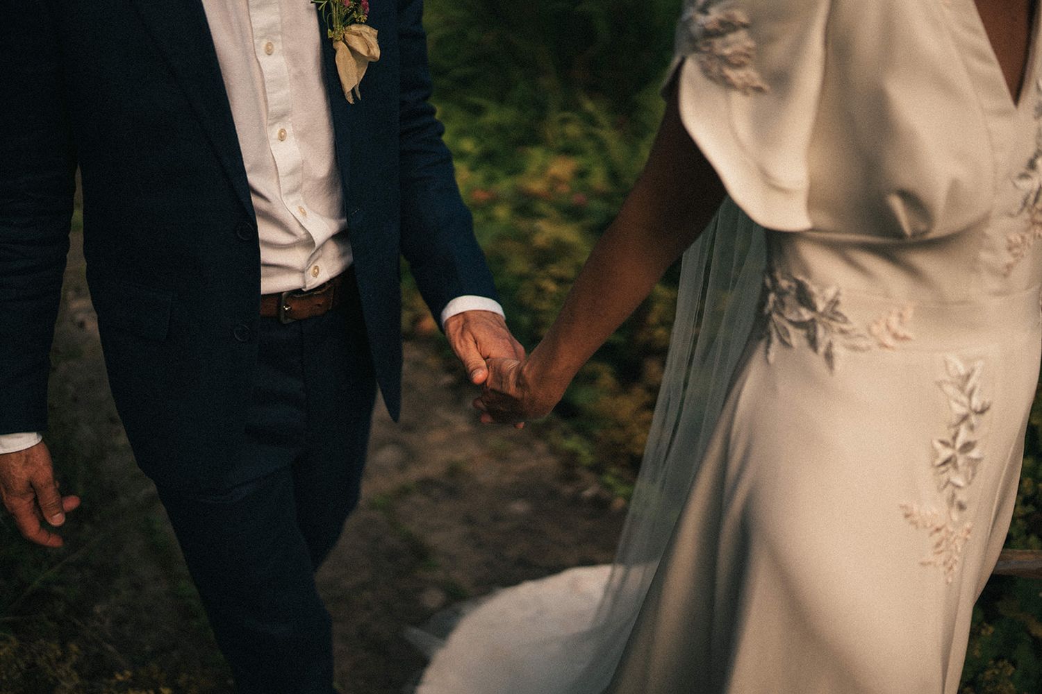 Close-up shot of a couple in formal attire holding hands outdoors with a focus on their interlocked fingers.
