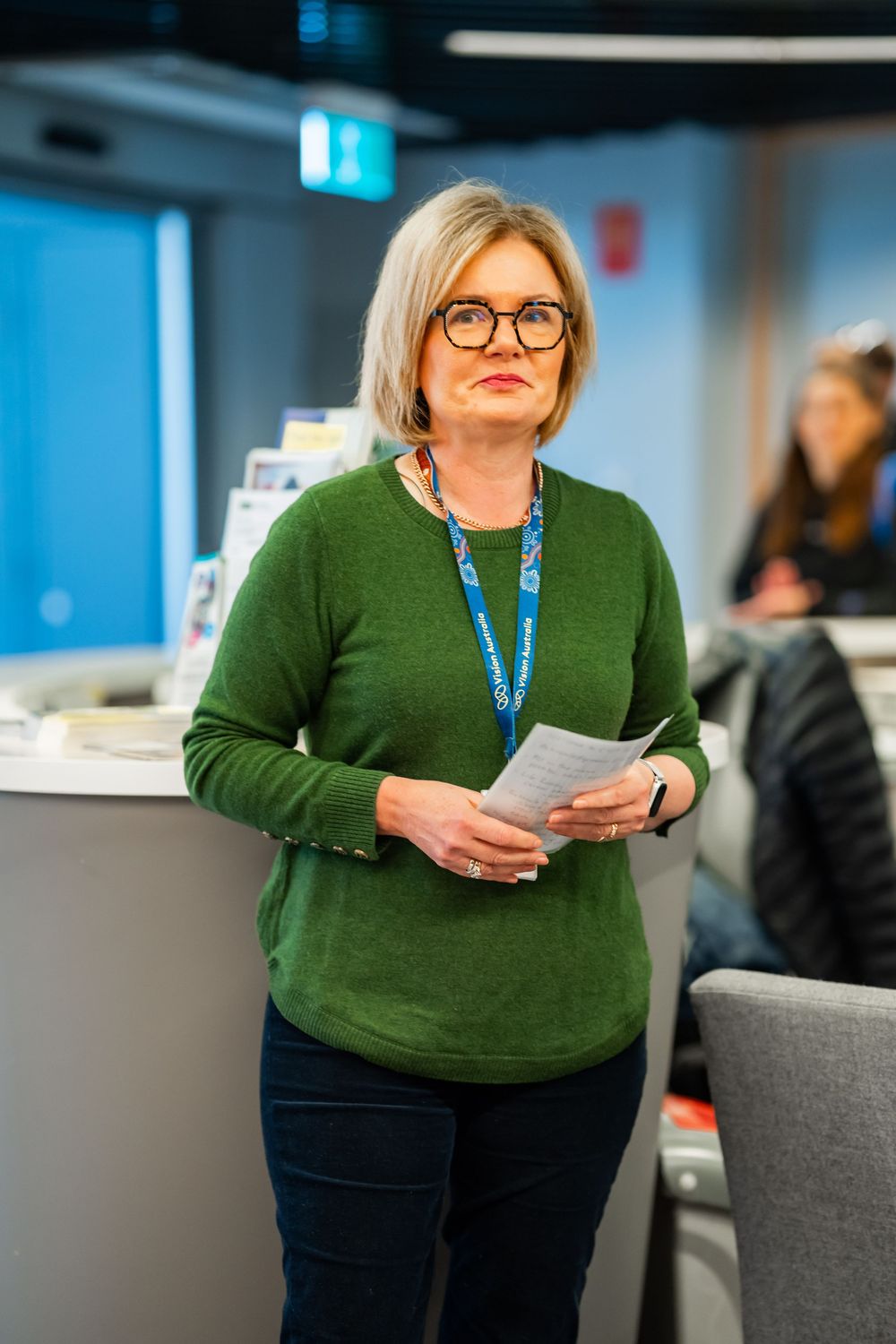 Professional wearing glasses and a green sweater stands in an office setting holding papers with a lanyard.