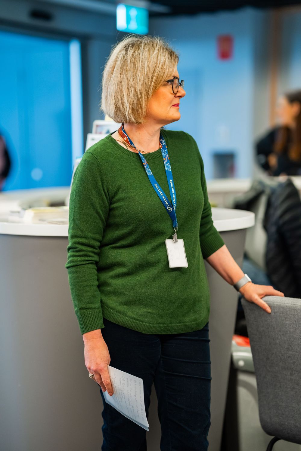 Professional wearing a green sweater and lanyard with ID badge stands in an office setting holding documents.