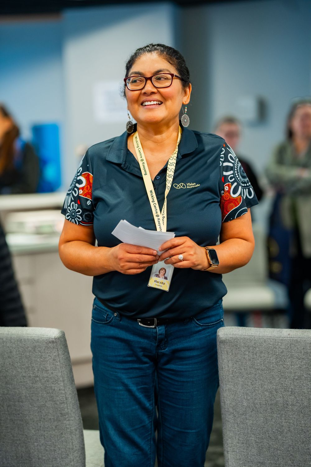 Professional wearing glasses, lanyard and navy polo with patterned sleeves stands with papers in an office setting.