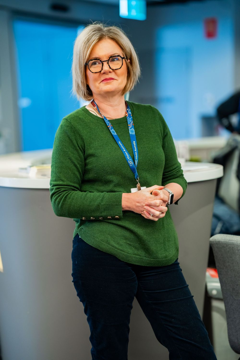 A professional wearing glasses and a green sweater stands in an office space with a lanyard and casual attire.
