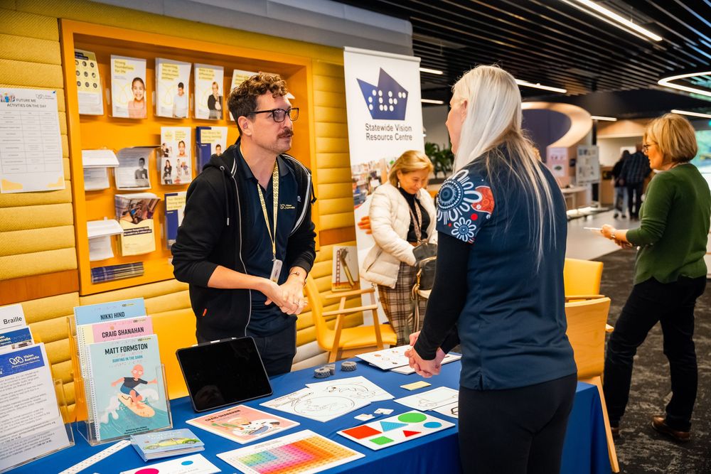 Two people talking at a career fair booth with informational materials and posters displayed on an orange wall behind them.