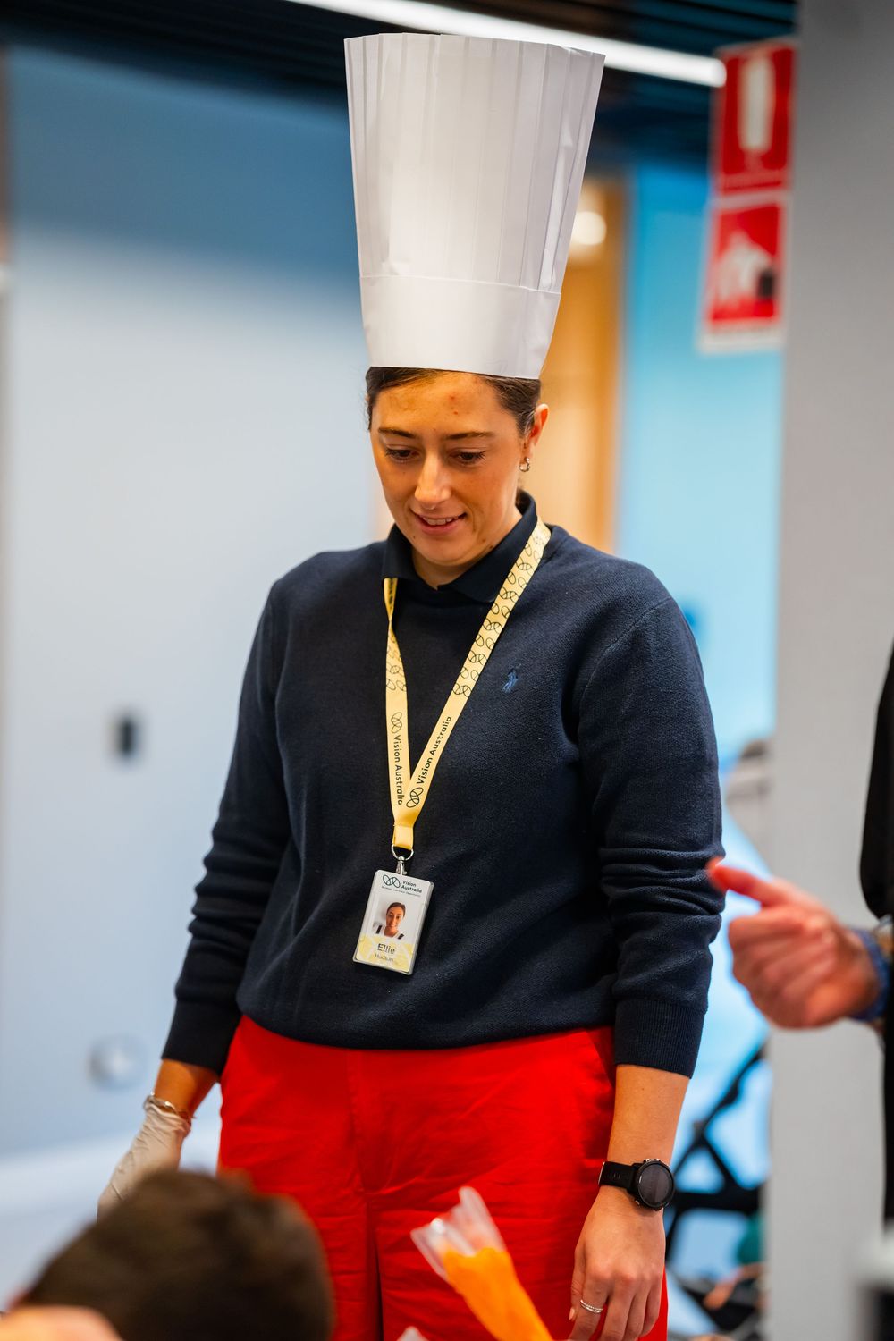 A chef in navy sweater and red pants wearing a tall white hat and ID badge smiles while working in a kitchen area.