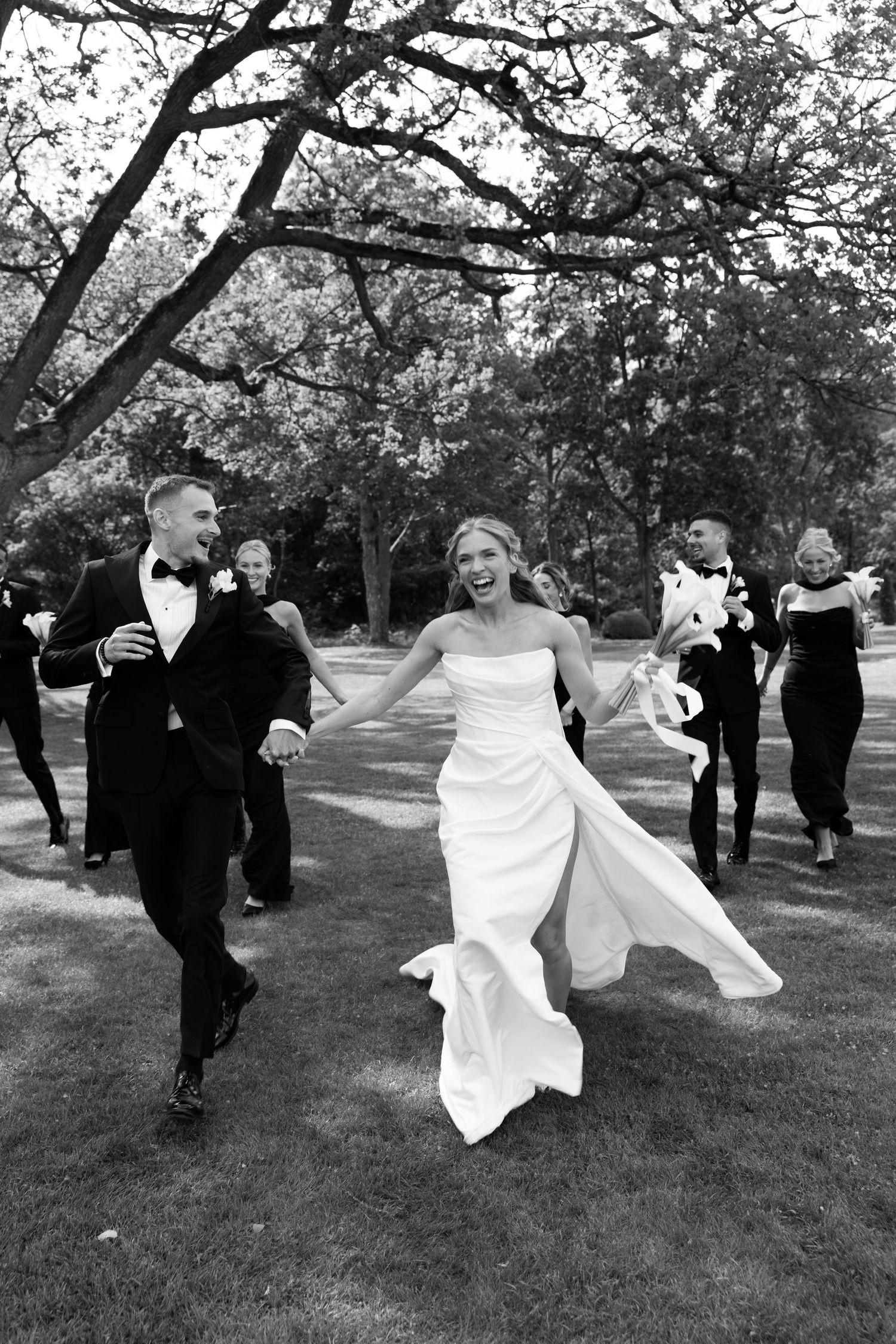 A joyful newlywed couple runs across a lawn with their wedding party following behind them in black attire.