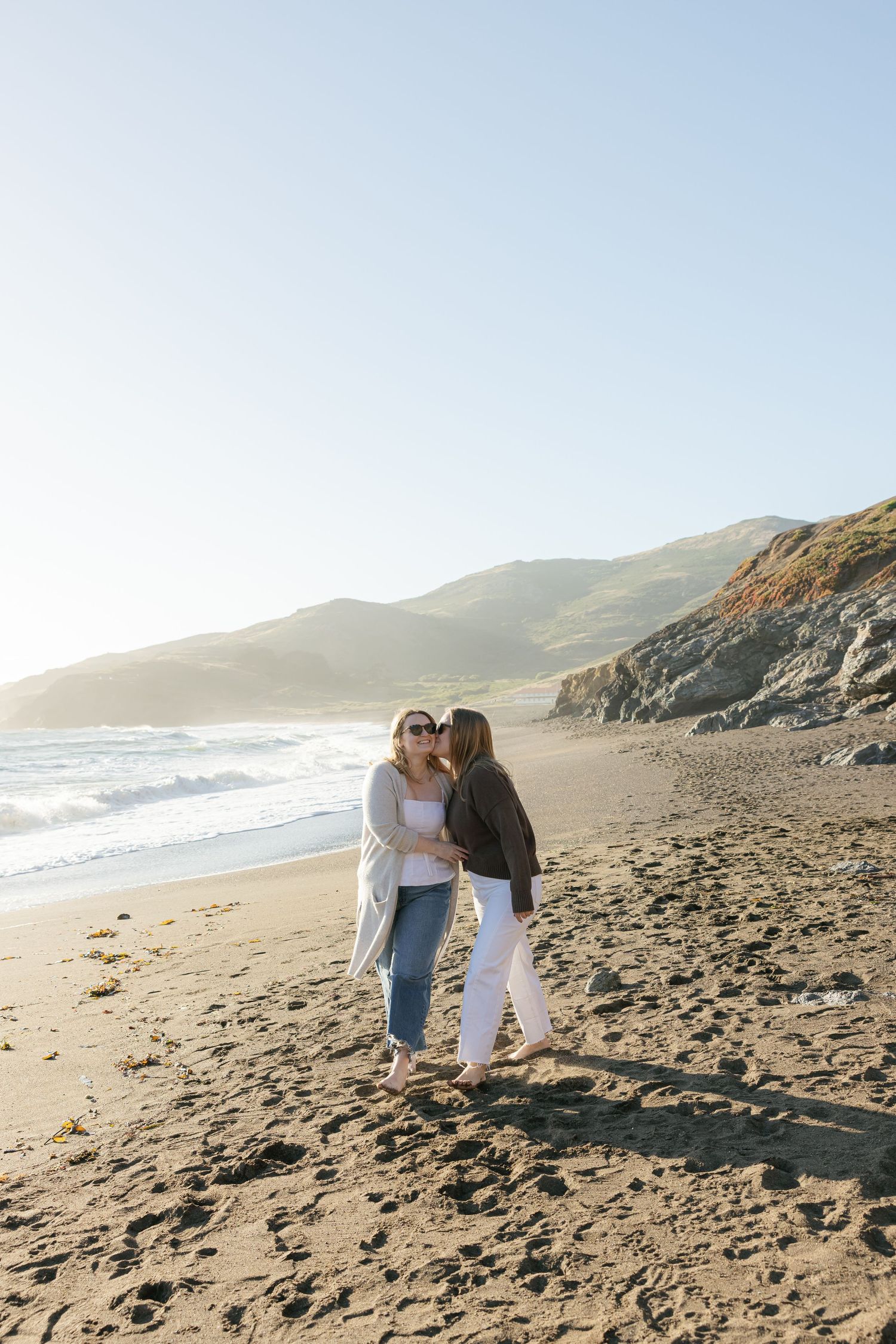 Two people share an intimate moment while walking along a scenic beach with rugged coastal cliffs at sunset.