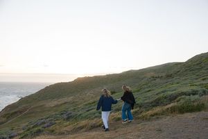 Two people walk along a scenic coastal trail at sunset with rolling green hills and ocean views.