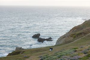 Rocky coastal cliffs overlook the blue Pacific Ocean with waves crashing against offshore rocks.