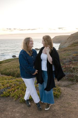 Two women share a lighthearted moment along a coastal cliff pathway at sunset with the ocean and rugged shoreline behind them.