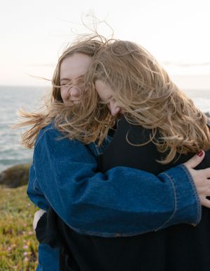 Two people with windswept blonde hair embrace on a coastal hilltop with ocean views at sunset.