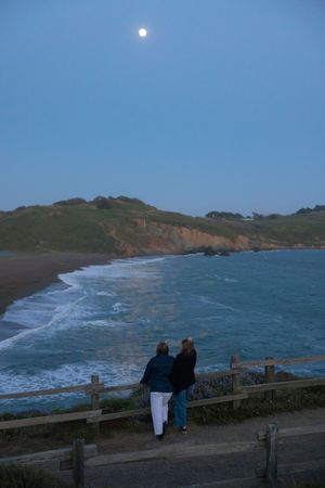 Two people stand at a coastal overlook watching waves crash on the beach under a full moon at dusk.