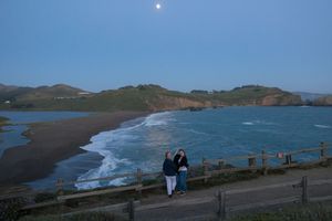 Two people admiring the moon over a scenic coastal beach view at dusk.