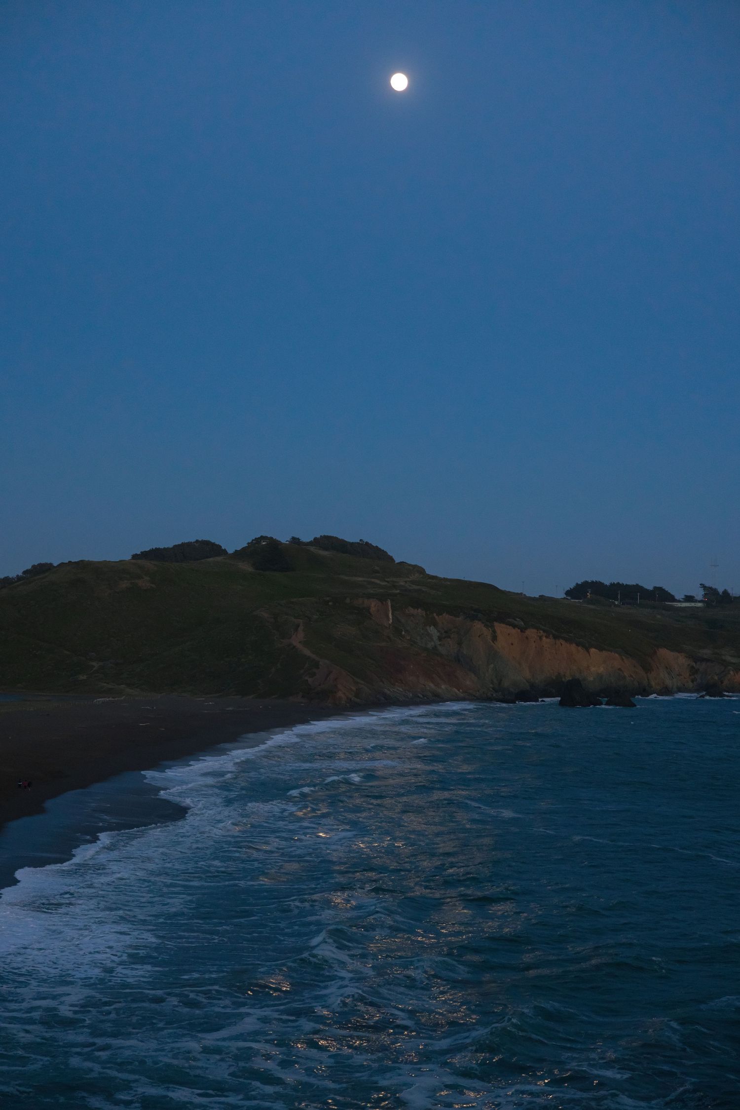 Full moon rises above a rugged coastal cliff with crashing waves meeting dark beach at twilight.