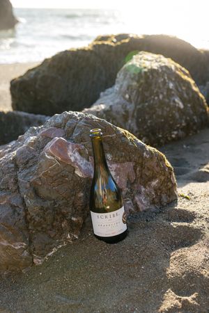 A wine bottle of Schild Estate wine sits among large rocks on a sandy beach with ocean waves in the background.